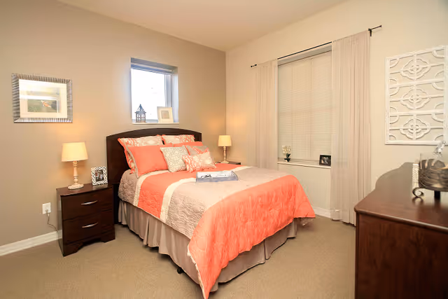 A neatly made bed with coral and beige bedding in a bedroom. The room has beige walls, a small window above the bed, two bedside tables with lamps, a framed picture on the wall, and a dresser on the right side. Sheer curtains cover a larger window on the right wall.