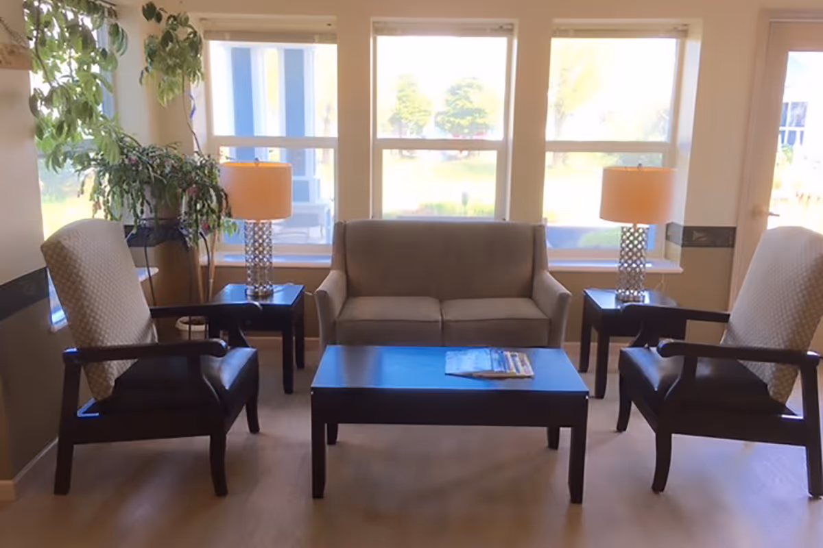 Sunlit sitting area with a sofa, two armchairs, a coffee table, side tables with lamps, and a potted plant in front of large windows.
