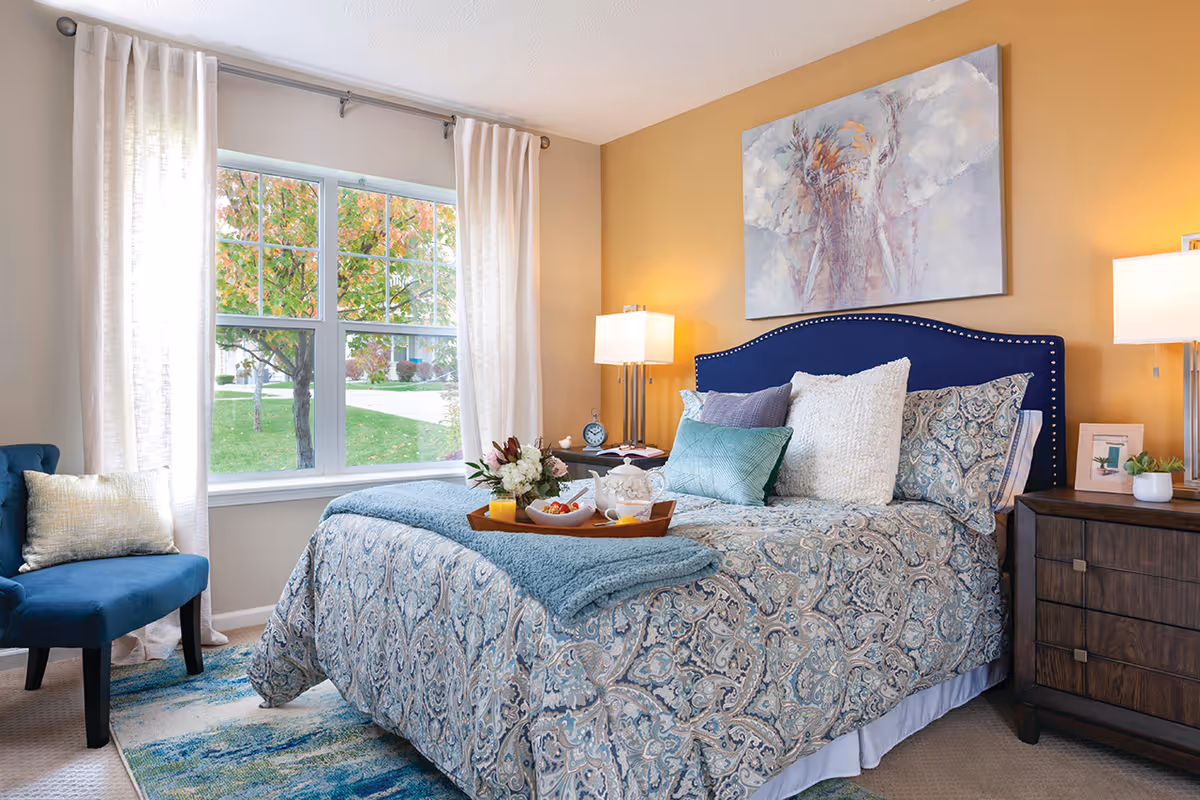 A cozy bedroom with a large window showing autumn trees outside. The bed has a blue patterned comforter, multiple pillows, and a blue upholstered headboard. There is a wooden nightstand with a lamp, a small plant, and a framed photo. A blue chair with a light-colored pillow is placed near the window. A painting of an elephant hangs above the bed.