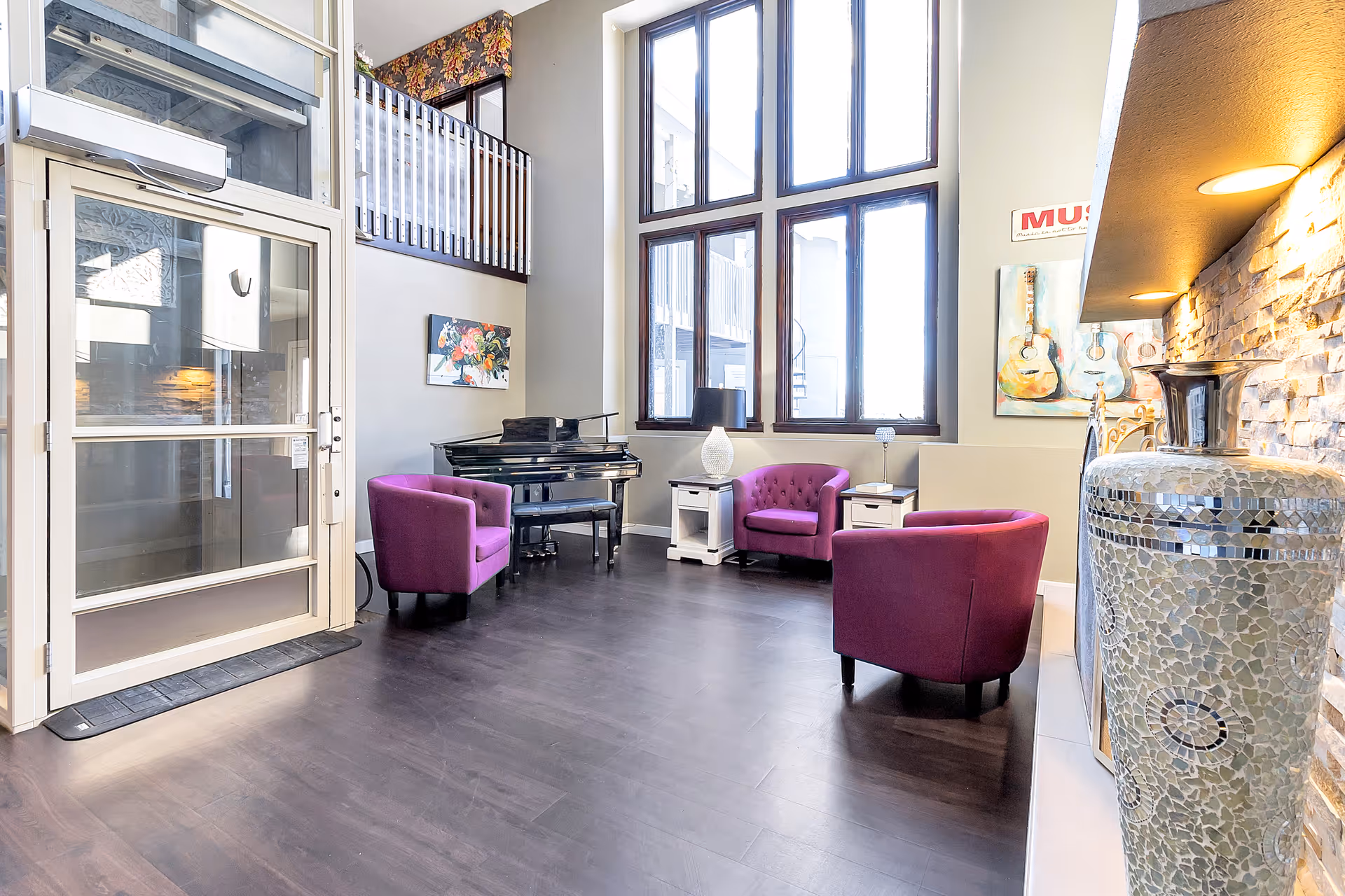 A bright and spacious senior living common area with large windows letting in natural light. The room features three purple armchairs arranged around two white side tables with lamps. A black grand piano is positioned against the wall beneath a floral painting. The floor is dark wood, and there is a large decorative mosaic vase near a stone accent wall with built-in lighting. A glass door leads outside, and a balcony railing is visible above.