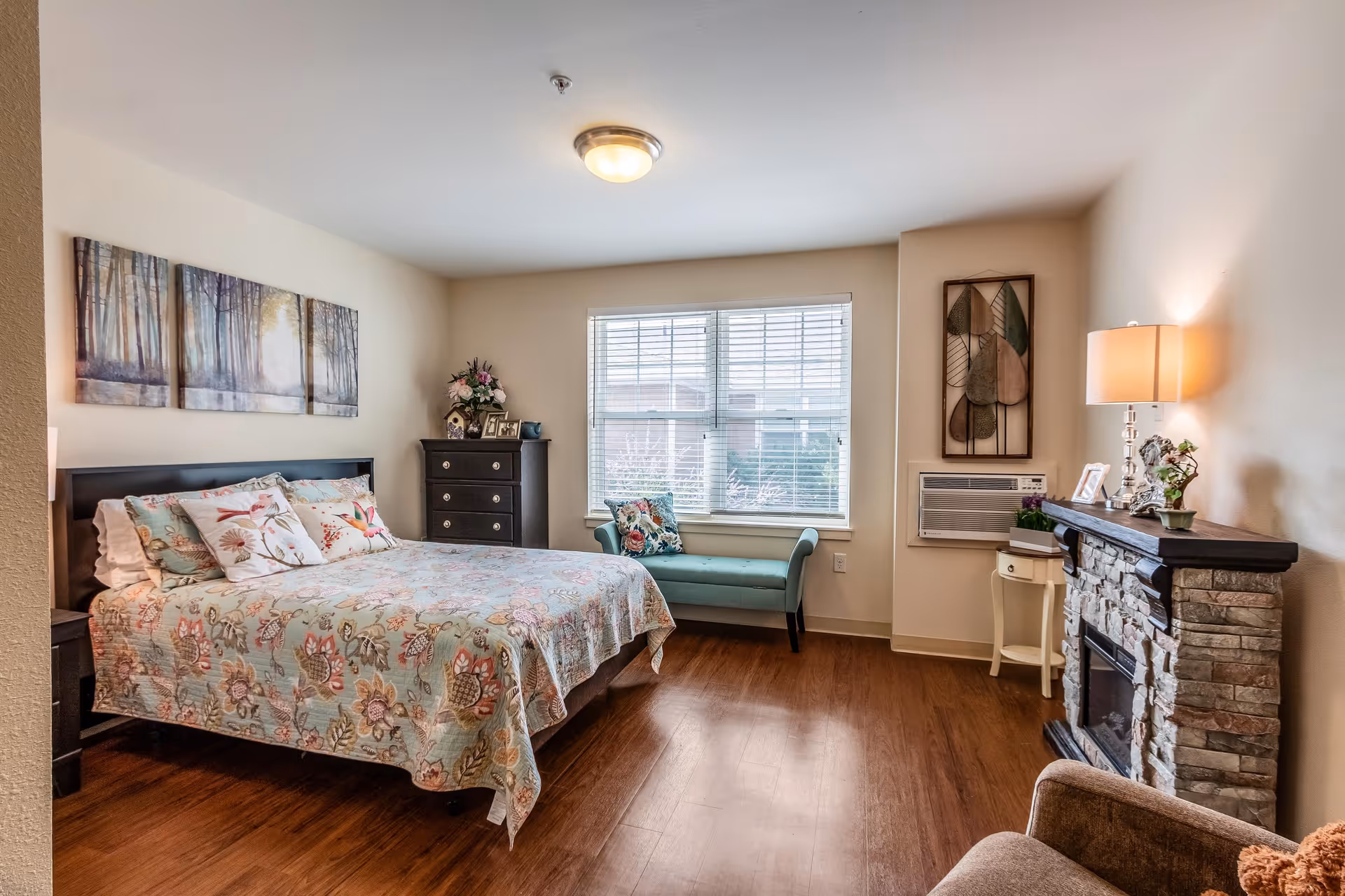 A cozy bedroom with a floral bedspread on a double bed, a dark wood dresser with framed photos and flowers, a teal upholstered bench under a window with blinds, a stone fireplace with a lamp and decorative items on the mantel, and a comfortable armchair in the corner.