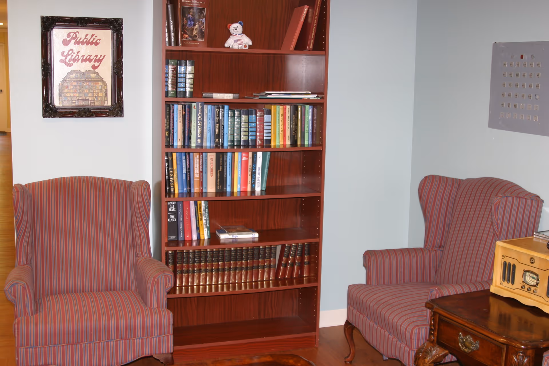 A cozy reading nook with two red and gray striped wingback chairs facing a wooden bookshelf filled with books and a small teddy bear on the top shelf. A framed sign on the wall reads 'Public Library'. A wooden side table with a vintage radio sits next to one of the chairs.