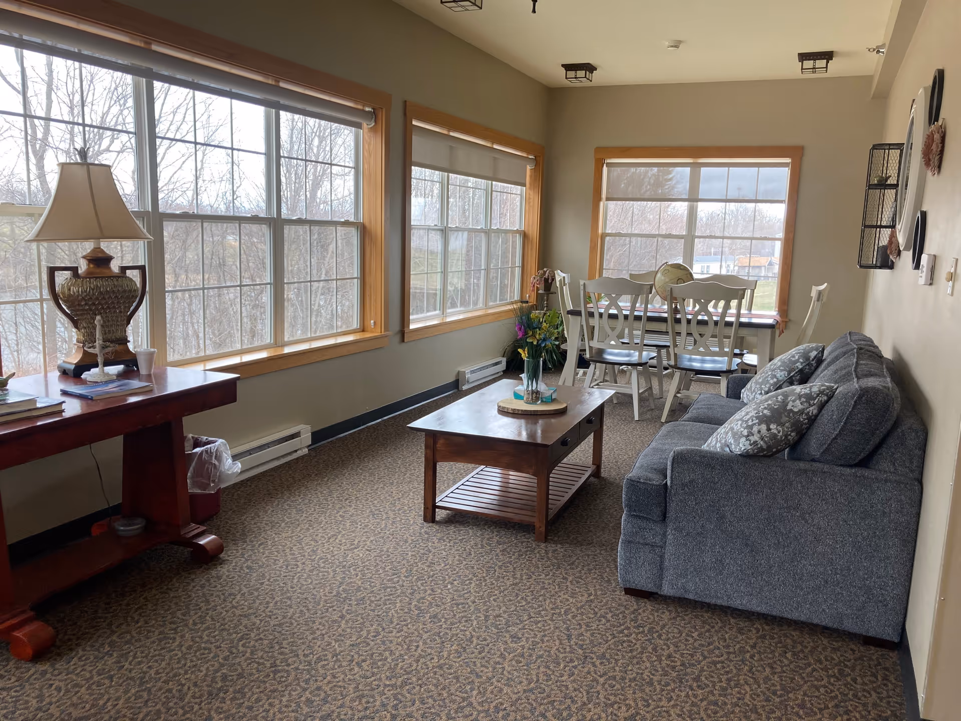 A bright living room area with large windows showing a view of trees outside. The room features a gray couch with patterned pillows, a wooden coffee table with a flower vase, a wooden side table with a lamp, and a dining table with four chairs near the windows.