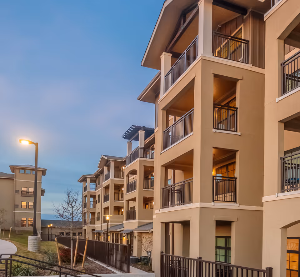 Exterior view of a multi-story senior living community building at dusk with balconies, railings, and lit street lamps along a paved walkway.