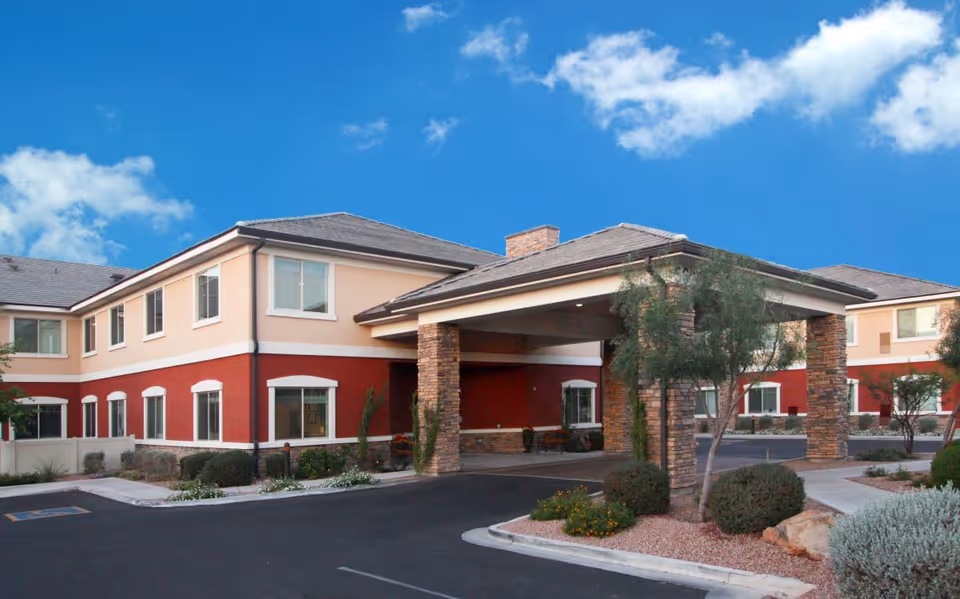 Exterior view of Legacy House of Mesa, a two-story building with beige and red walls, a covered entrance supported by stone pillars, surrounded by landscaped bushes and trees under a blue sky with some clouds.