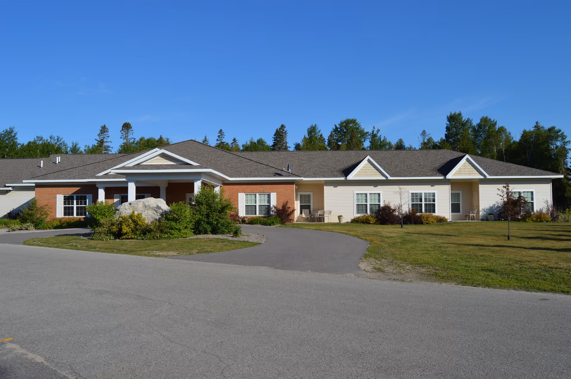 Front exterior view of Cedar Cove Assisted Living facility, a single-story building with a combination of brick and siding, surrounded by a driveway, lawn, and trees under a clear blue sky.