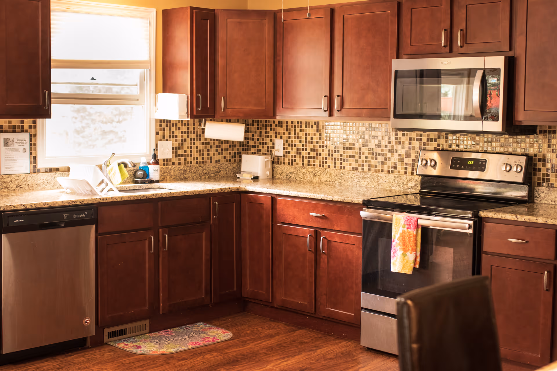 A kitchen with wooden cabinets, a granite countertop, a mosaic tile backsplash, a stainless steel dishwasher, stove, and microwave. There is a window above the sink with a dish rack and cleaning supplies on the counter. A colorful towel hangs on the oven handle and a small rug is on the wooden floor.