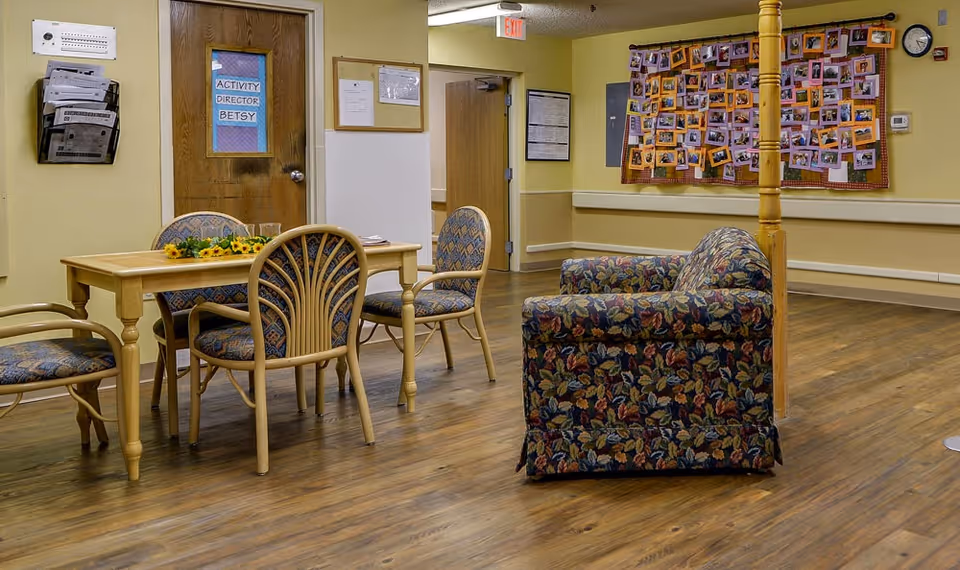 A nursing home common room with a table and chairs, a patterned armchair, and a photo-filled bulletin board on the wall.