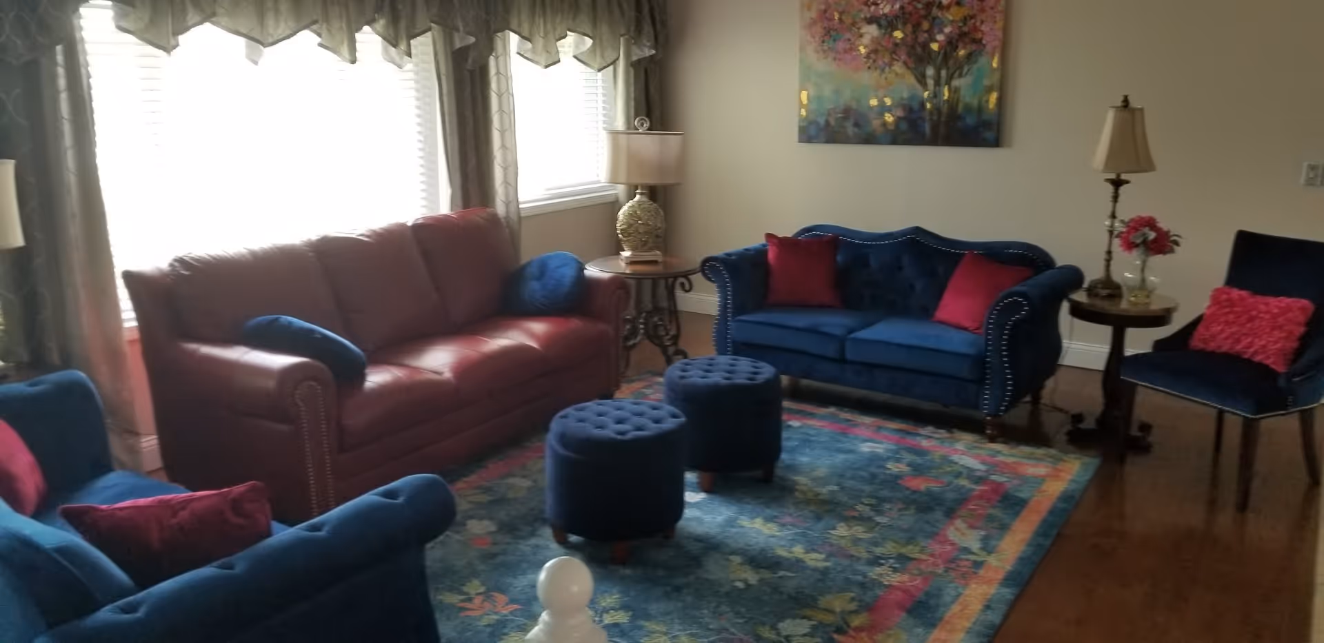 A cozy living room with a red leather sofa, two blue velvet sofas, and a blue velvet chair. The sofas have red and blue cushions. There are two round blue ottomans on a colorful floral area rug. Two side tables with lamps and a vase with flowers are placed near the sofas. A colorful painting hangs on the wall above one of the blue sofas. Large windows with curtains allow natural light into the room.