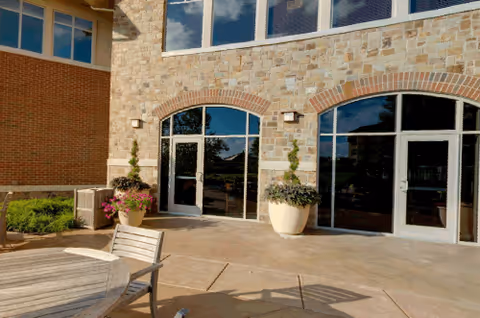Outdoor patio area with a round wooden table and chairs in front of a building with large arched glass doors and stone walls. Two large potted plants flank the doors, and the sky is partly cloudy.