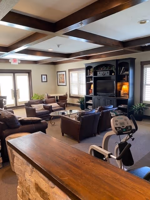A cozy living room area with brown leather chairs and sofas arranged around a glass coffee table. There is a large dark wooden entertainment center with a TV and shelves holding books and decorative items. The room has wooden ceiling beams, windows with white shutters, and a stationary exercise bike in the foreground. A wooden counter with a stone base is also visible.