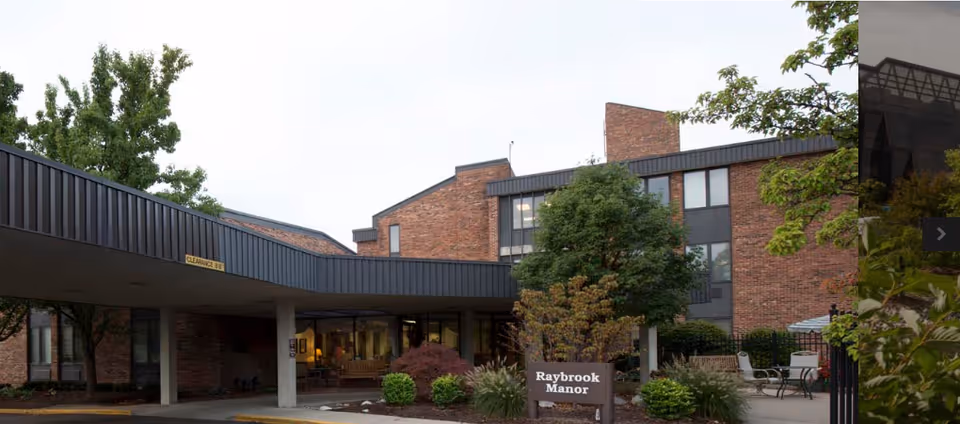 Front exterior of a brick senior living building with a covered entrance and a sign reading 'Raybrook Manor'.