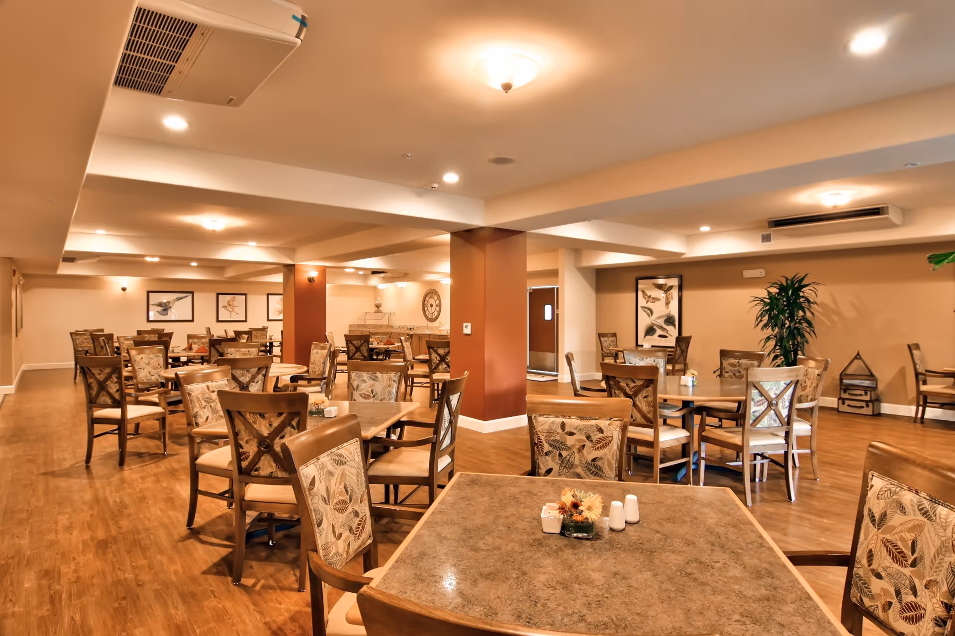 A spacious dining room with multiple wooden tables and chairs featuring leaf-patterned upholstery. The room has warm lighting, wooden flooring, and beige walls decorated with framed bird artwork and a large clock. There are plants and decorative items placed around the room.