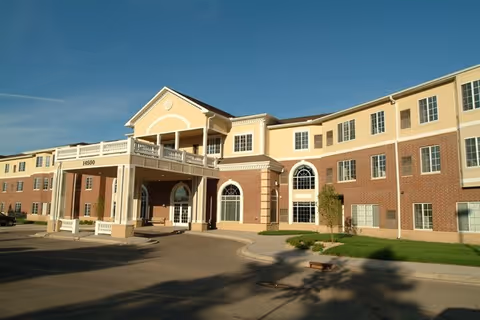 Exterior view of a three-story senior living facility building with a covered entrance and columns, under a clear blue sky.