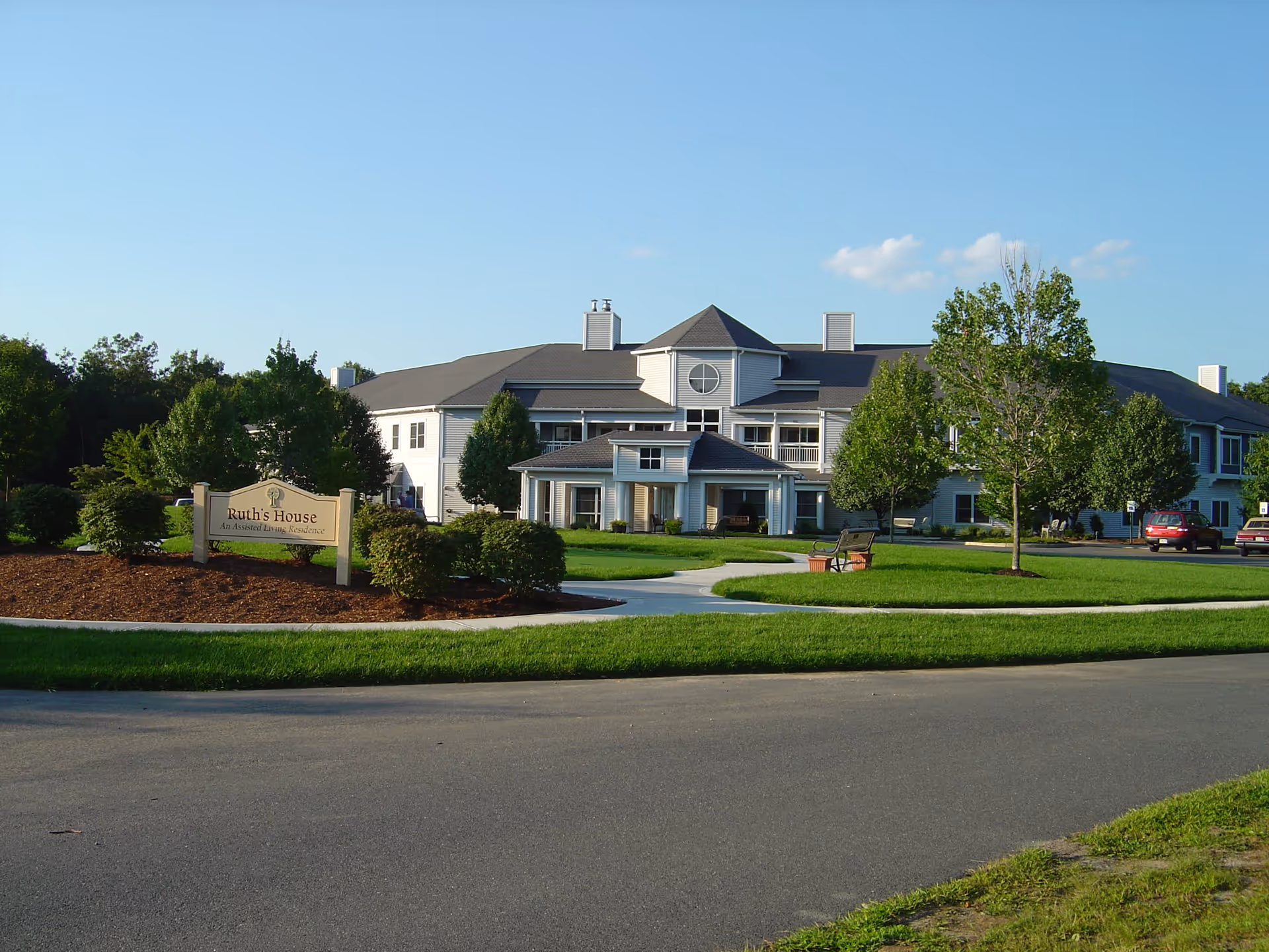 Front view of a two-story assisted living building with a manicured lawn, walkway, benches, and a sign reading "Ruth's House".