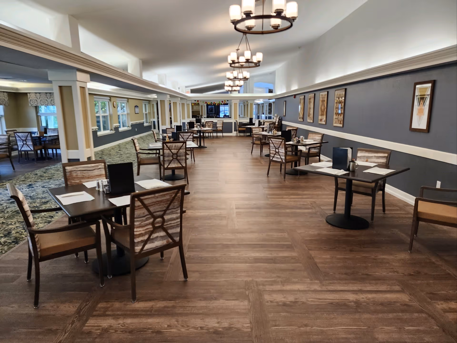 A spacious dining room in a senior living facility with multiple tables and chairs arranged neatly. The room features wooden flooring, modern chandeliers hanging from the ceiling, and framed artwork on the walls. Large windows allow natural light to brighten the space.