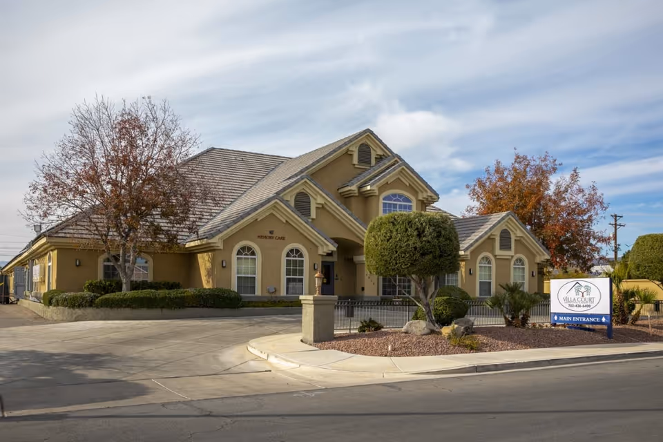 Front exterior of the Villa Court Assisted Living building with trimmed landscaping, trees, and a driveway entrance sign.