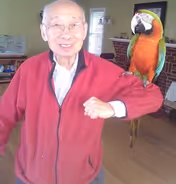 An elderly man wearing glasses and a red jacket is smiling indoors with a colorful parrot perched on his arm. The background shows a living room with wooden floors, a fireplace, and some furniture.