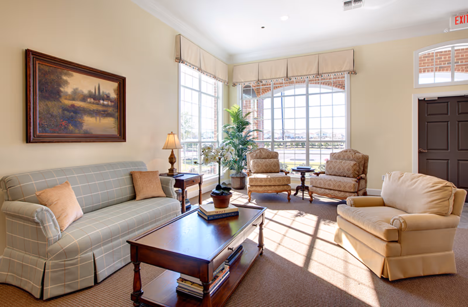 A bright and cozy living room with a large window letting in natural light. The room features a plaid sofa with two beige pillows, a wooden coffee table with books and a potted orchid, two patterned armchairs, a beige armchair, a side table with a lamp, a large potted plant, and a framed landscape painting on the wall.