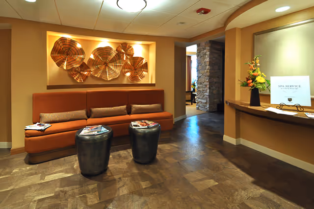 Reception lounge with an orange upholstered bench, two round stools, decorative metal wall art, and a reception counter with flowers.