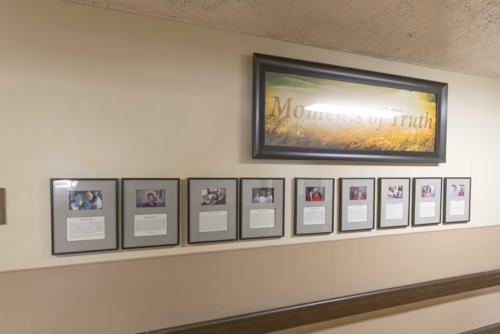 A hallway wall displaying a series of framed photographs and written stories beneath a larger framed picture with the words 'Moments of Truth'. The wall is painted beige with a darker beige wainscoting and a wooden handrail below the frames.
