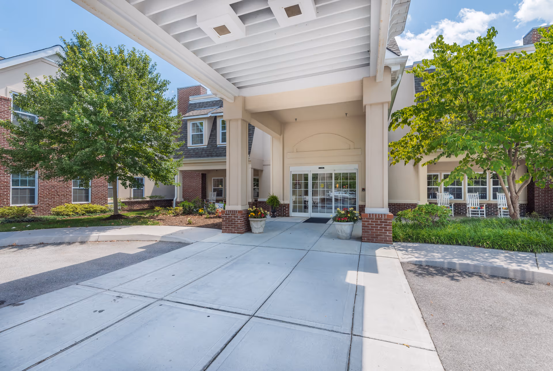 Entrance of Park Place of West Knoxville facility showing a covered driveway with two large columns, potted plants, and automatic glass doors. The building exterior features brick and beige siding with windows and greenery including trees and shrubs on either side.