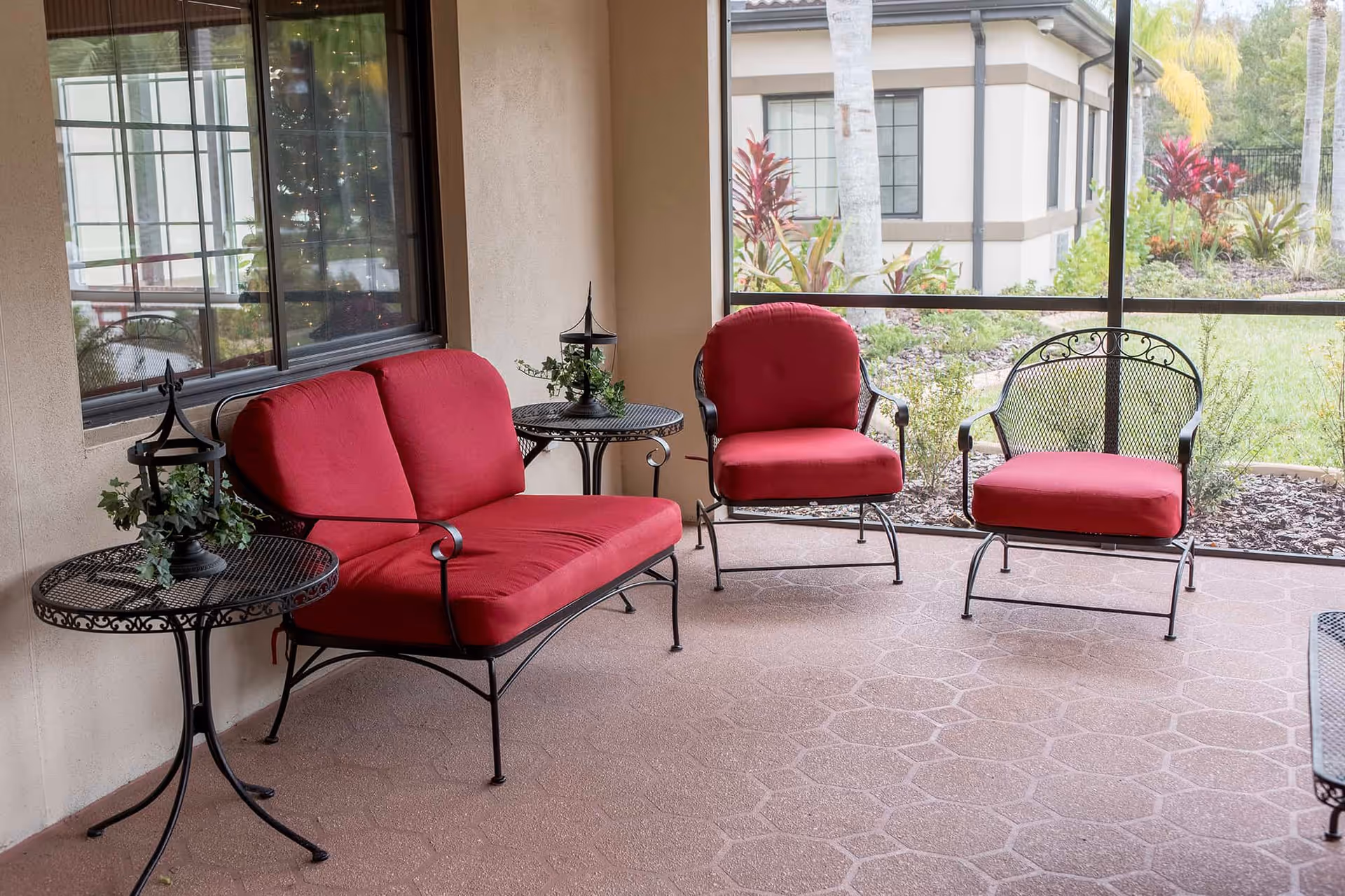 Screened outdoor patio with red-cushioned metal loveseat and chairs arranged around small wrought-iron tables overlooking landscaped grounds.