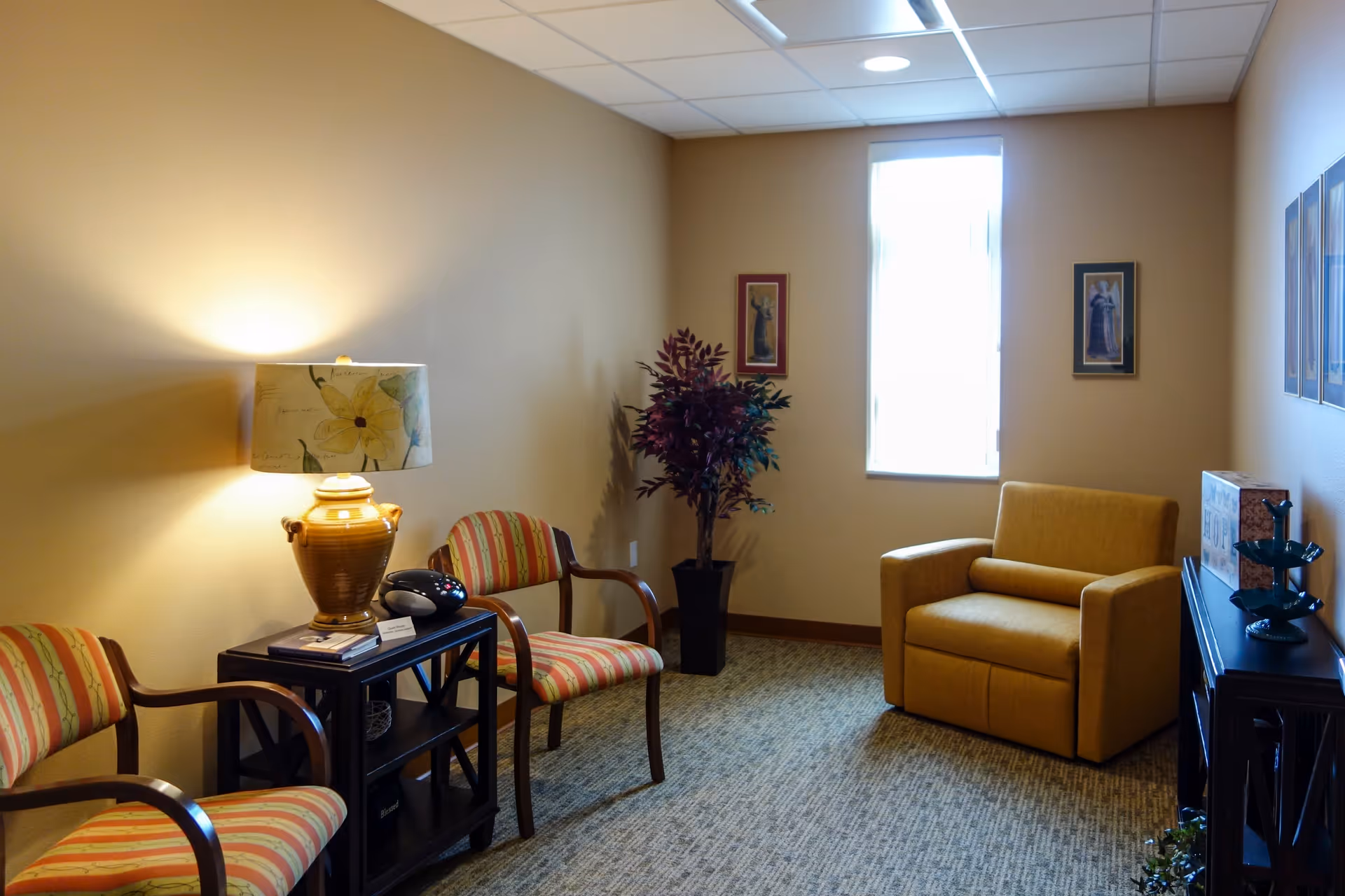 A cozy sitting area in a senior living facility with two striped armchairs, a small black side table with a decorative lamp and a phone, a yellow armchair, a tall potted plant, and framed artwork on beige walls with a window letting in natural light.