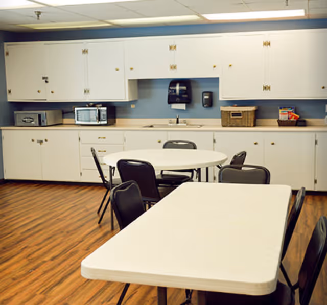 Communal dining area with folding tables and chairs in front of a kitchenette with white cabinets and a microwave.