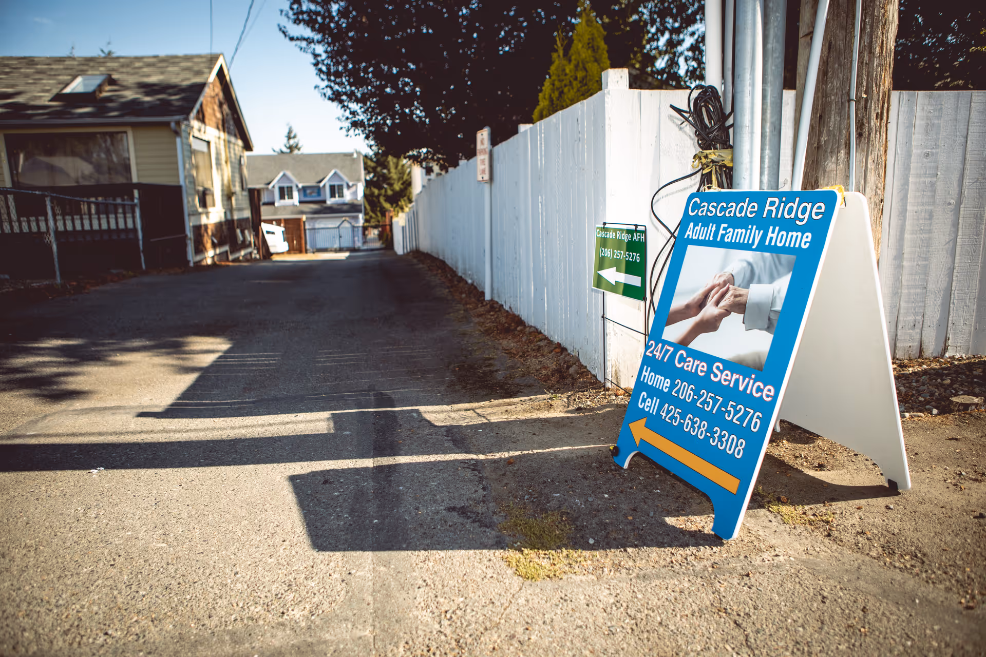 A sidewalk sign for Cascade Ridge Adult Family Home placed on a paved path next to a white fence. The sign displays contact information and an image of two people holding hands. Residential houses and trees are visible in the background under a clear sky.