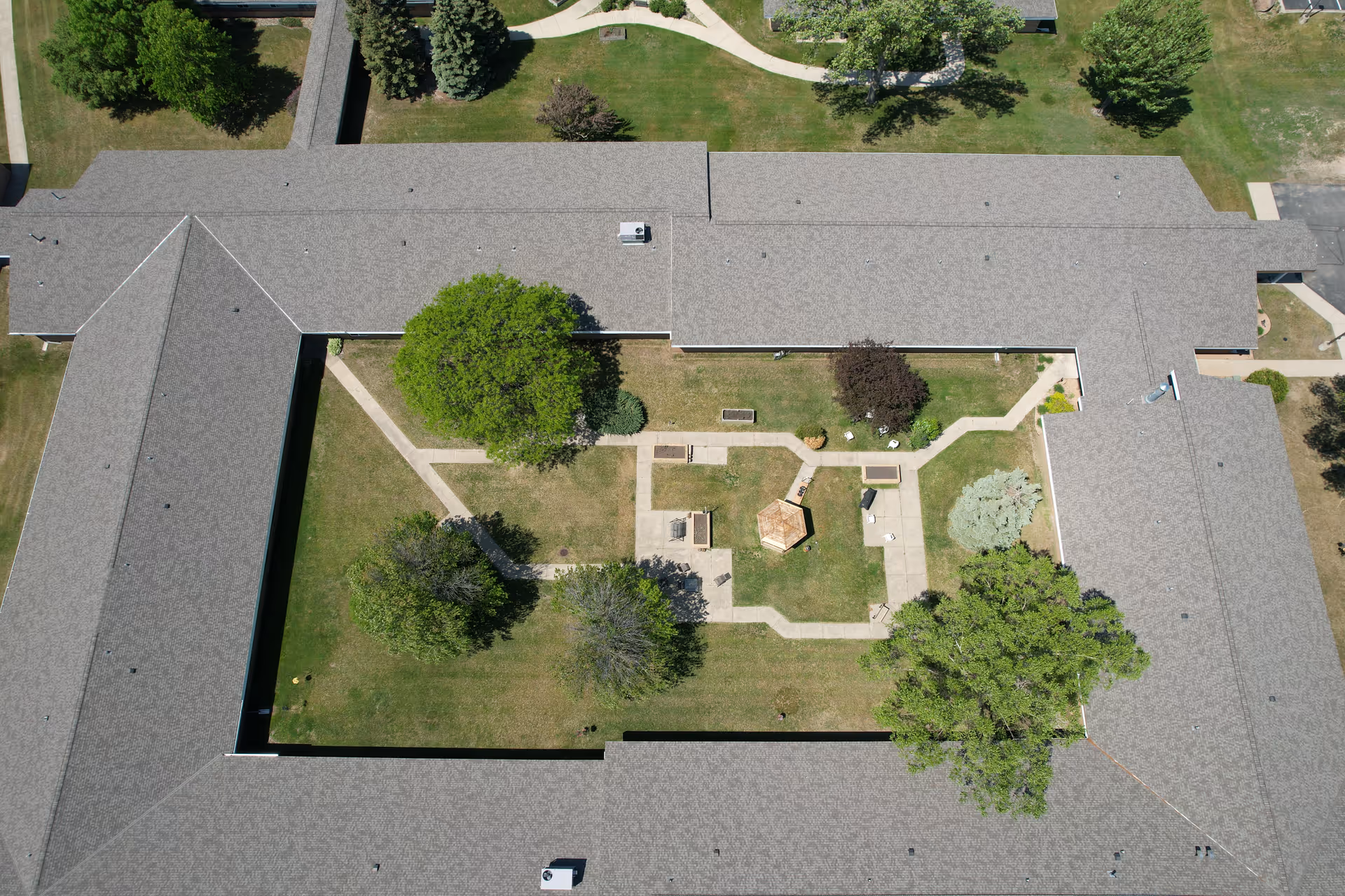 Aerial view of a senior living facility courtyard with a central gazebo, surrounded by walking paths, trees, and benches. The courtyard is enclosed by a building with a gray roof.