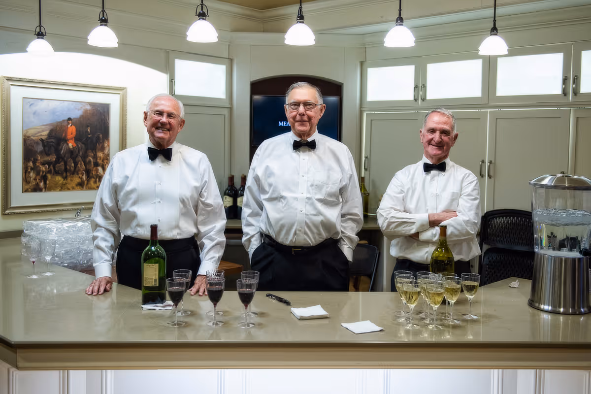 Three elderly men in white shirts and black bow ties stand behind a counter with wine glasses and bottles in a well-lit indoor bar area.