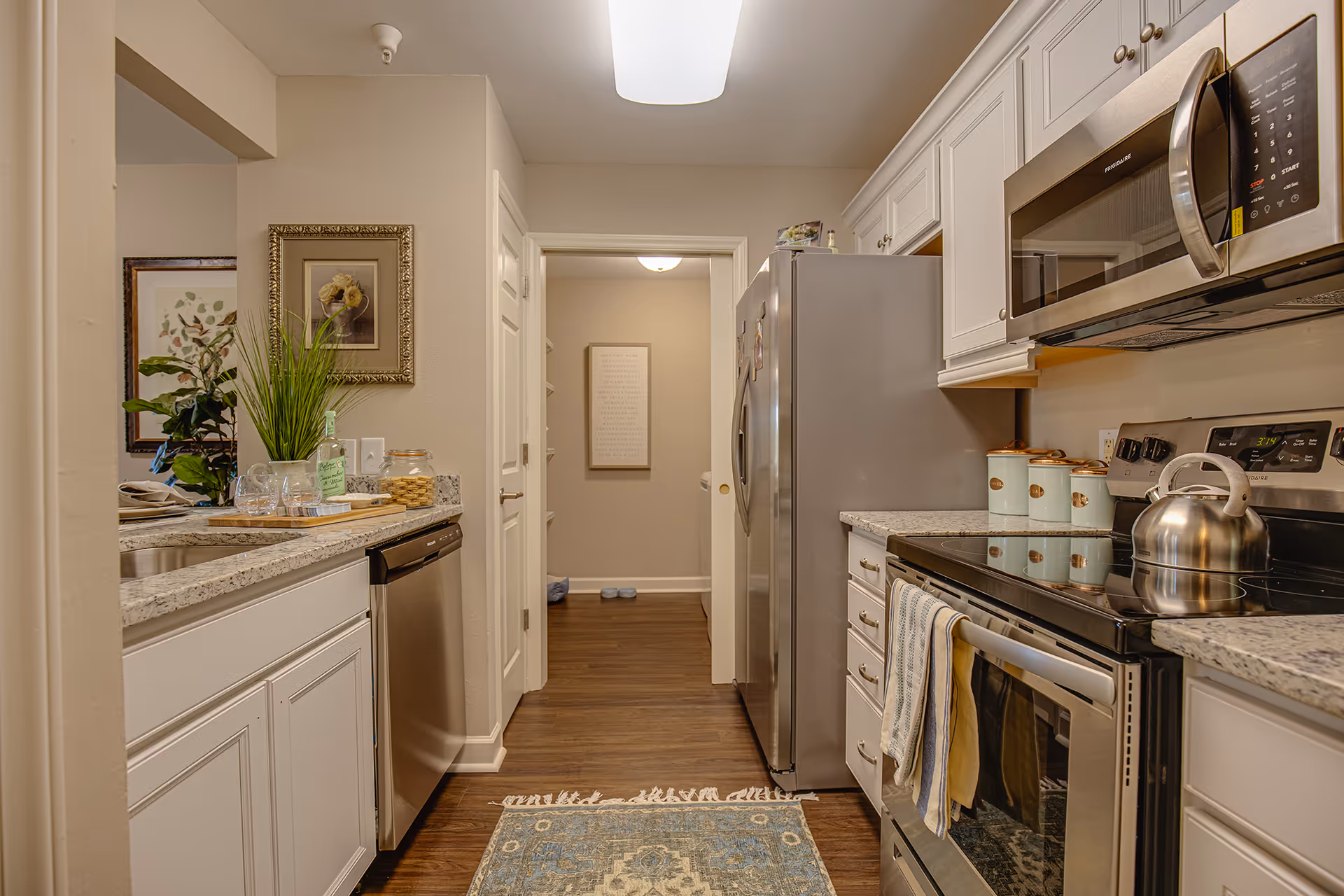 A modern kitchen with white cabinets, granite countertops, and stainless steel appliances including a refrigerator, dishwasher, microwave, and electric stove with a kettle on it. There is a small blue patterned rug on the wooden floor, a plant and framed artwork on the left wall, and a hallway visible in the background.