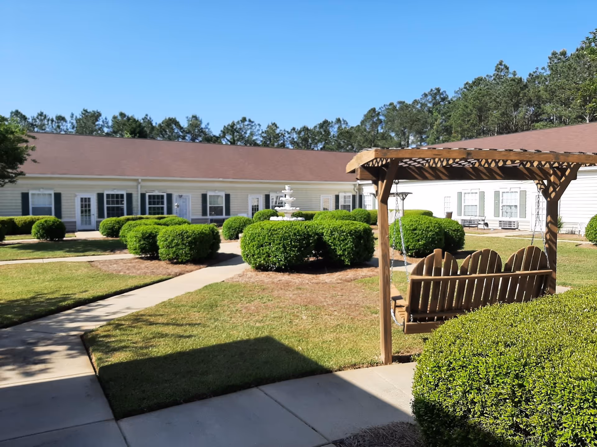 Outdoor courtyard area of a senior living facility with neatly trimmed bushes, a concrete walkway, a wooden swing under a pergola, and a white multi-tiered fountain in the center. The building surrounding the courtyard has white siding, multiple windows with shutters, and several doors. Trees are visible in the background under a clear blue sky.