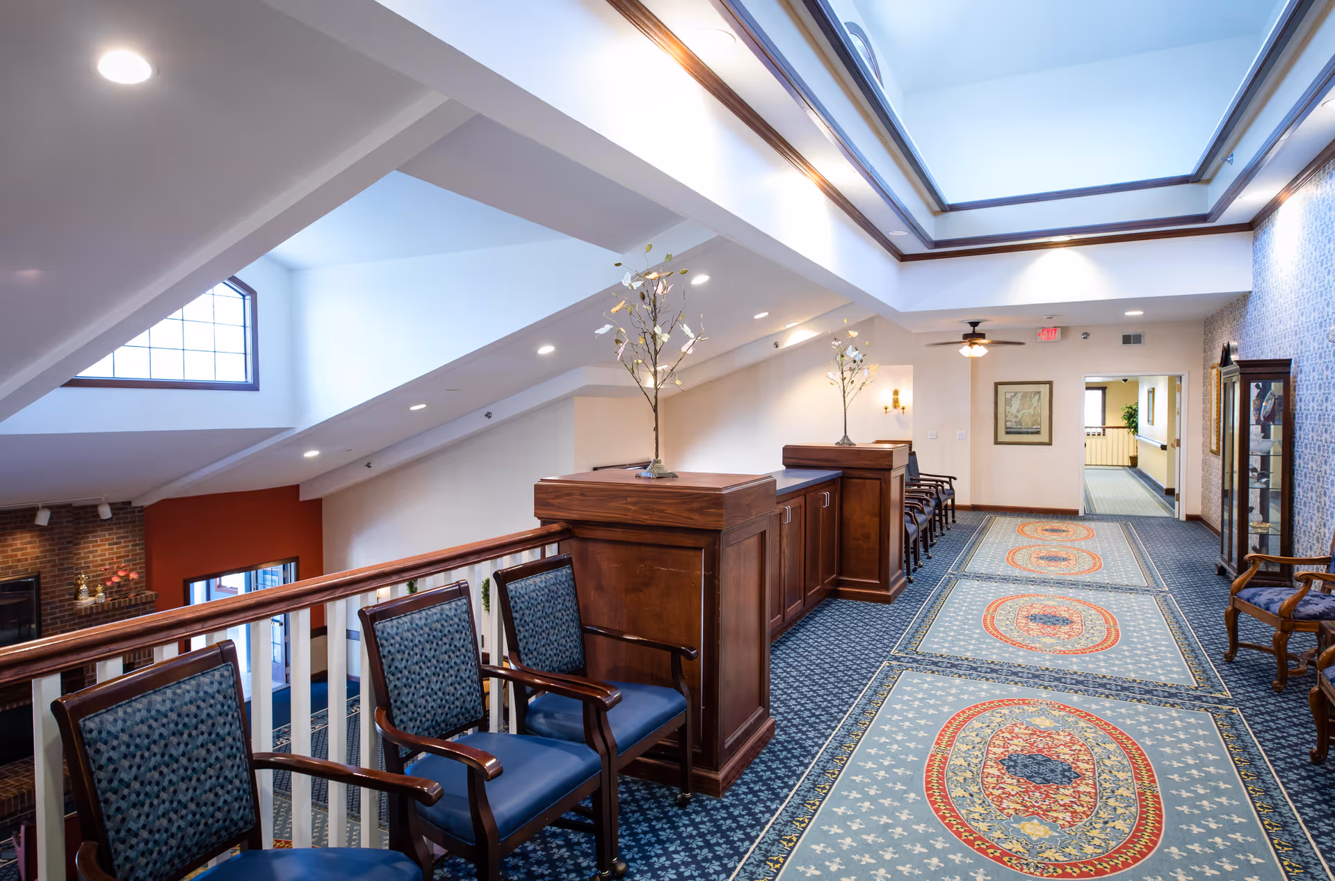 Upstairs atrium hallway in an assisted living facility with seating, wooden railings, skylights, and patterned carpet.