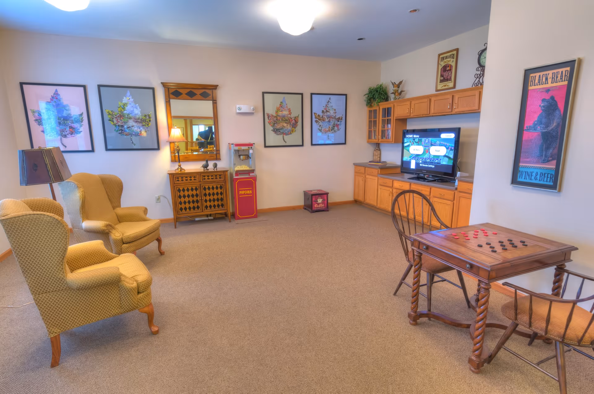A bright common room with upholstered wingback chairs, a game table, a popcorn machine, framed art, and a TV with built-in cabinetry.