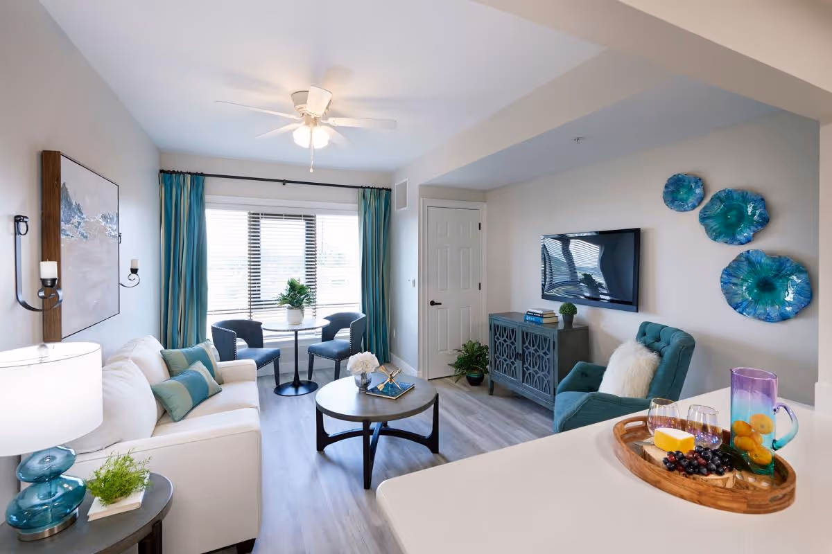 A bright and modern living room with a white sofa adorned with teal and green pillows, a round coffee table with decorative items, two blue chairs around a small round table near a window with teal curtains, a teal armchair with a white fluffy pillow, a wall-mounted flat-screen TV above a decorative cabinet, and three blue-green decorative wall plates. A tray with a pitcher of infused water, glasses, cheese, and grapes is on a white countertop in the foreground.