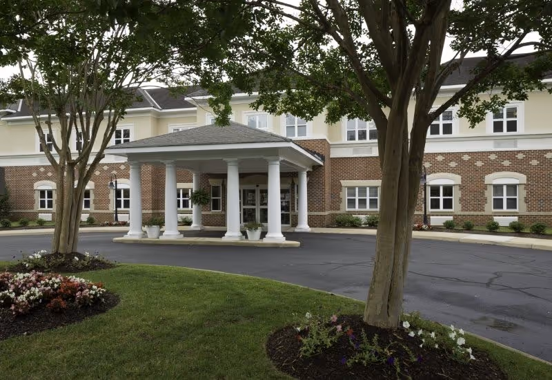 Front exterior view of a senior living facility named Harmony at Independence, featuring a covered entrance with white columns, a circular driveway, manicured lawns, flower beds, and trees.