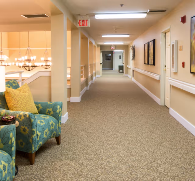 Carpeted corridor in a senior living facility with patterned armchairs, handrails, and an exit sign.
