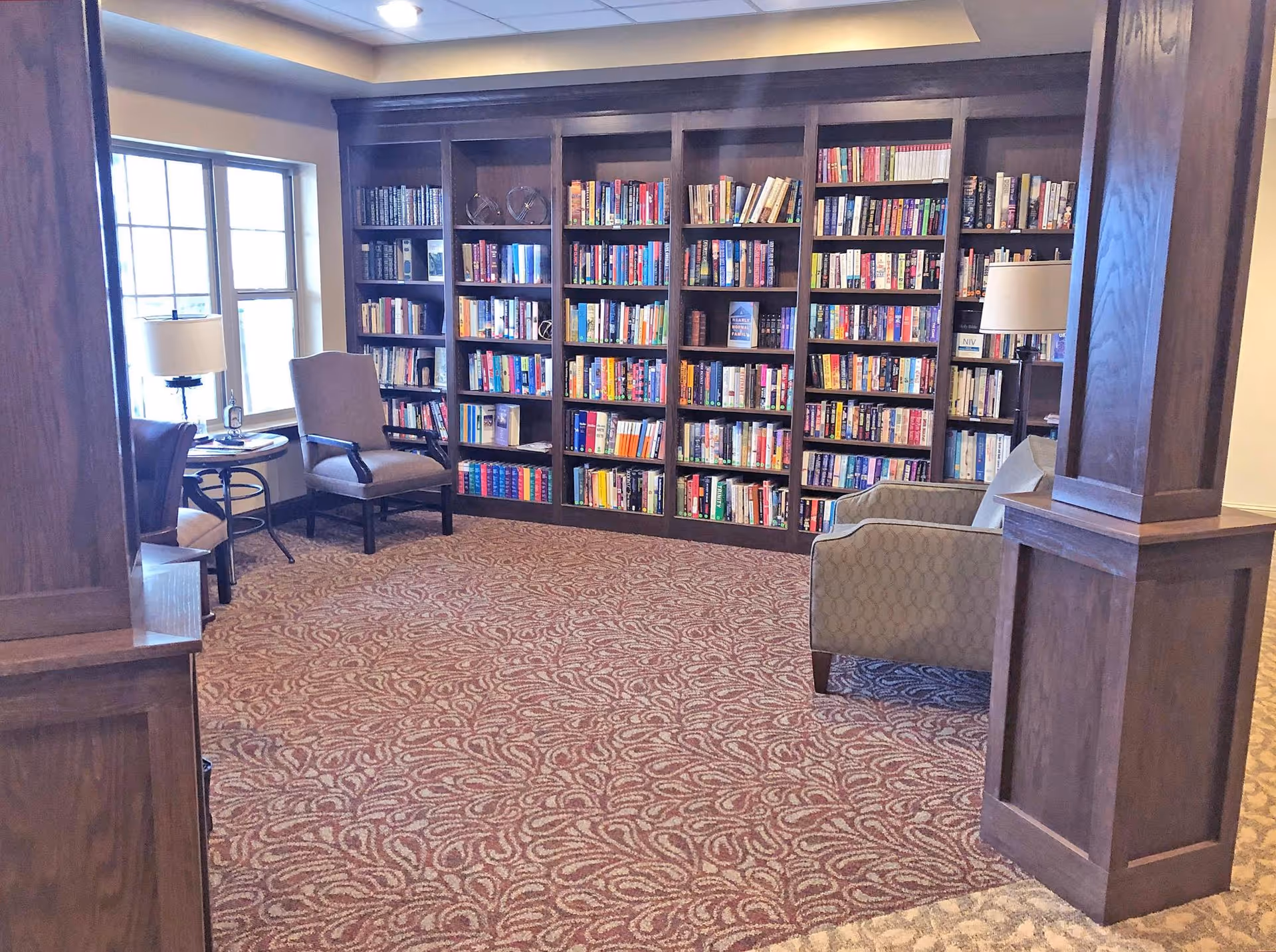 A cozy library room with large wooden bookshelves filled with books along the back wall. There are several comfortable chairs and small tables near a window letting in natural light. The room has patterned carpet and wooden pillars.