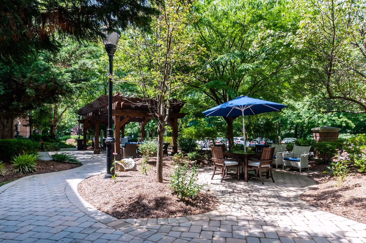 A peaceful outdoor garden area with a paved walkway, surrounded by trees and shrubs. There is a wooden pergola with seating underneath and a round table with four chairs and a blue umbrella providing shade. A lamppost is visible along the walkway.