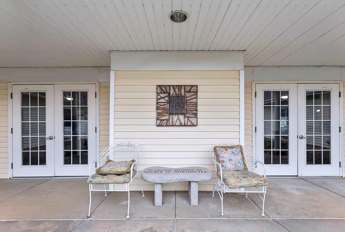 Covered outdoor seating area with two white metal chairs with floral cushions and a small stone table between them. The table has an inscription in memory of someone. Behind the seating area is a beige wall with a decorative clock mounted on it. There are two sets of white double doors with glass panes on either side of the seating area.