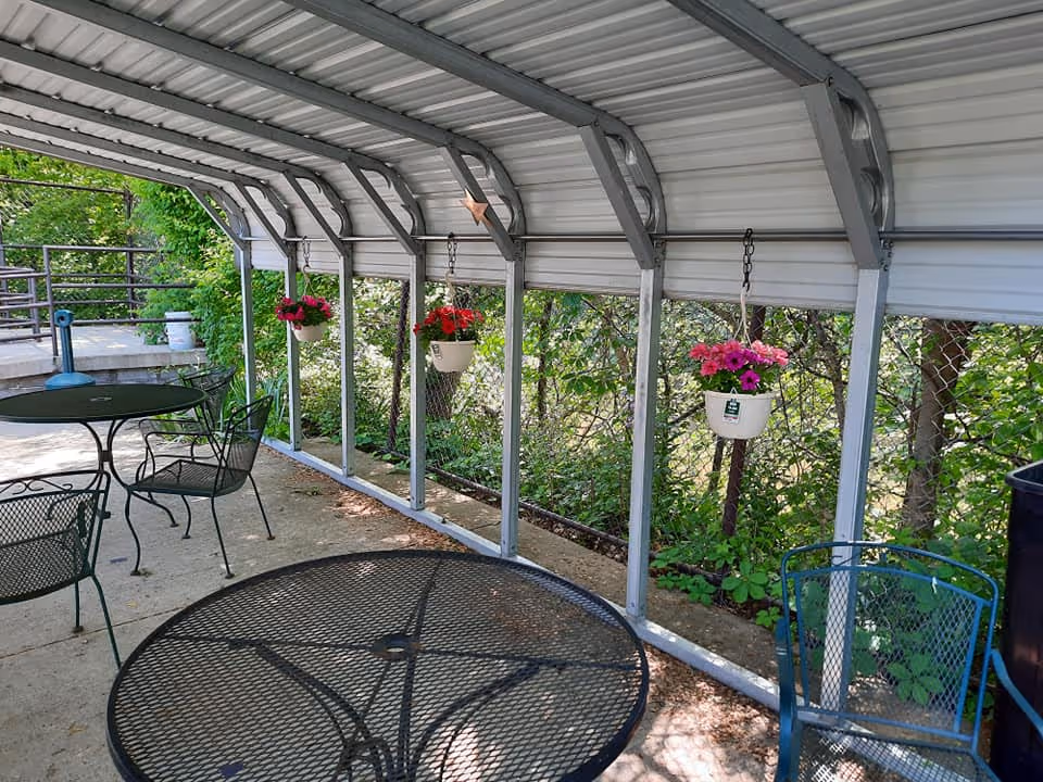 Covered outdoor patio area with metal mesh tables and chairs. Hanging flower pots with pink and red flowers are suspended from the metal roof structure. Green foliage and trees are visible beyond the patio.