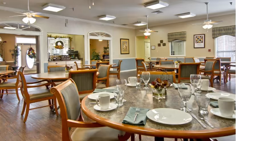 A dining room in Henley Place with multiple round tables set with white plates, cups, glasses, and silverware. The room has wooden chairs with green upholstery, hardwood floors, ceiling fans, and framed artwork on the walls. Large windows with valances allow natural light into the space.