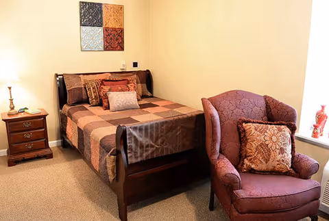 A cozy bedroom with a wooden bed featuring a patchwork quilt and multiple decorative pillows. Next to the bed is a wooden nightstand with a lamp and some items on it. A comfortable upholstered armchair with a floral cushion is placed near a window with a red vase on the windowsill. The walls are light-colored with a decorative wall hanging above the bed.