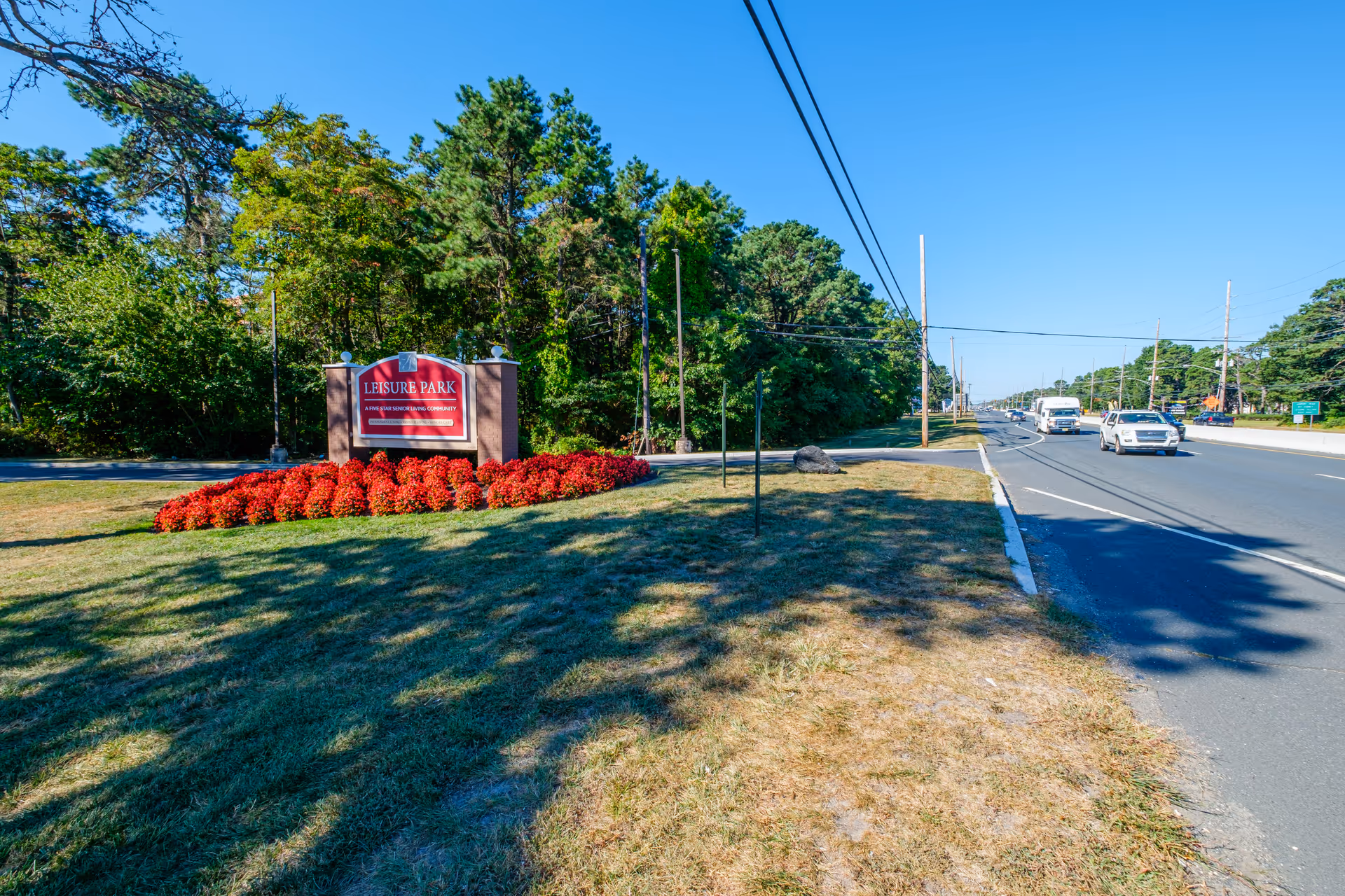 Entrance sign for Leisure Park on a grassy median with red flowers, trees, and a road with cars.
