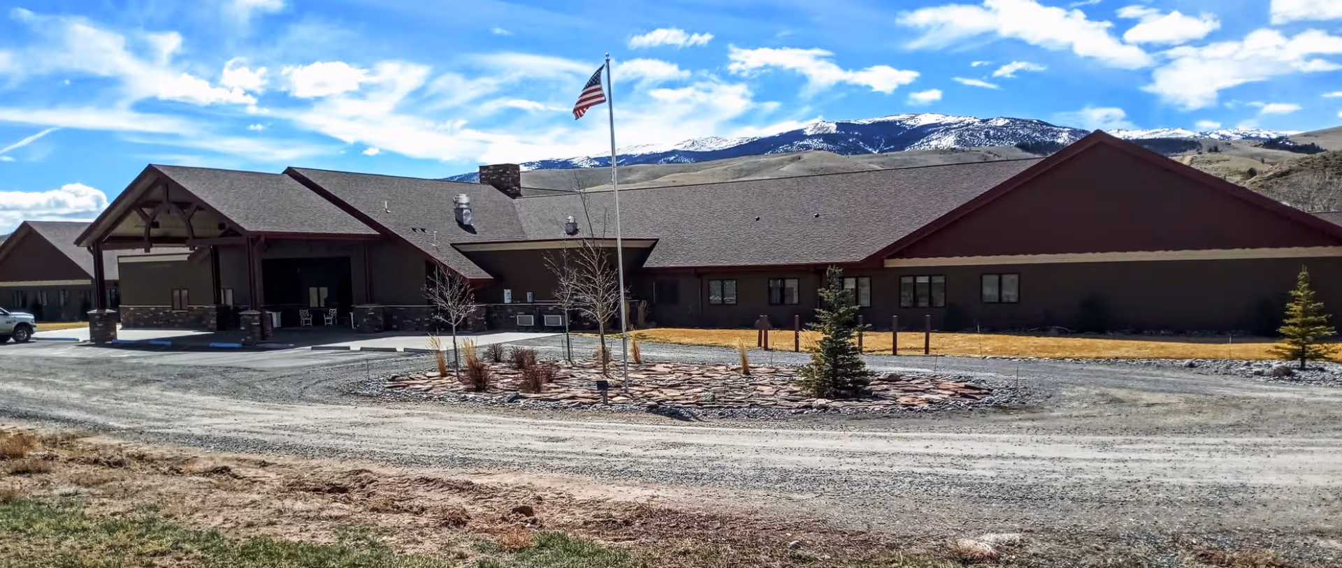 Exterior view of Warm Valley Lodge - Assisted Living, a single-story building with a pitched roof, an American flag on a flagpole in front, and a circular landscaped area with small trees and shrubs. Snow-capped mountains and a partly cloudy blue sky are visible in the background.