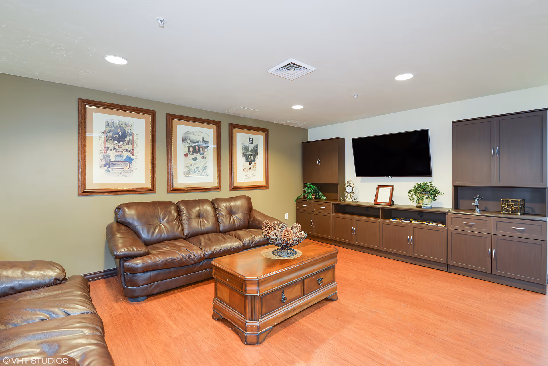 A cozy living room with brown leather sofas, a wooden coffee table with decorative pine cones, three framed pictures on the wall, and a large flat-screen TV mounted above dark wooden cabinets with plants and decorative items.