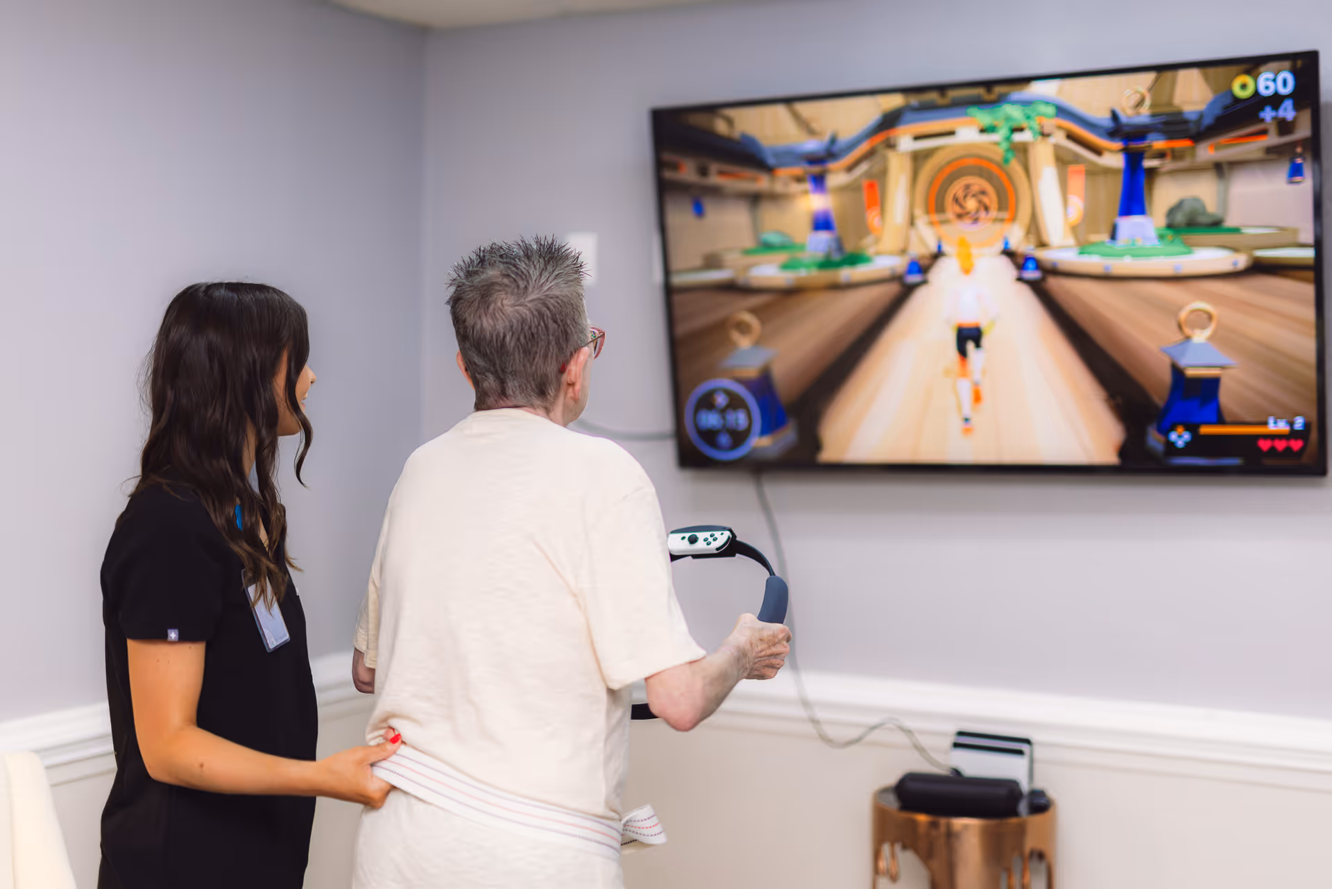An elderly person playing a video game using a motion controller while a caregiver stands beside them offering support in a room with light gray walls and a mounted flat-screen TV displaying the game.