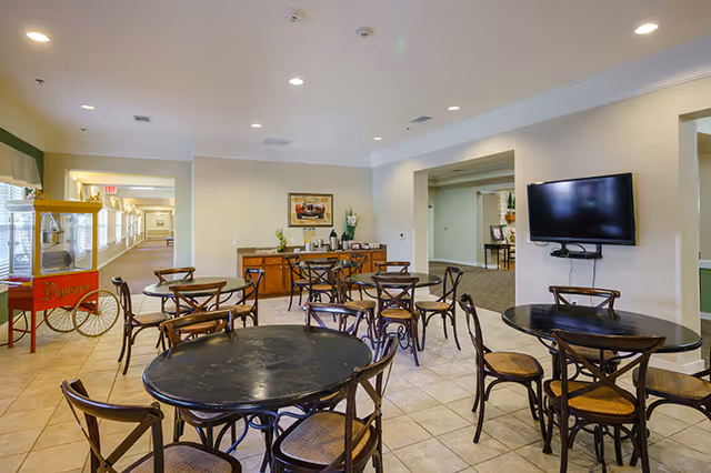 Interior view of a common area in an assisted living facility with several round tables and chairs arranged on a tiled floor. A popcorn machine is visible on the left side near a hallway. A wooden sideboard with decorative items and a coffee station is against the far wall. A flat-screen TV is mounted on the right wall. The space is well-lit with recessed ceiling lights.